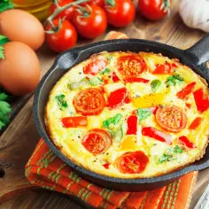 Frittata z warzywami w żeliwnej patelni, obok jajka, pomidory i natka pietruszki.