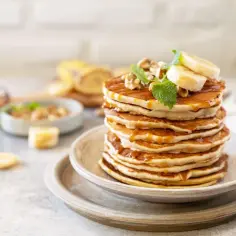Stos placków bananowych polanych syropem, ozdobiony bananem, orzechami i miętą.