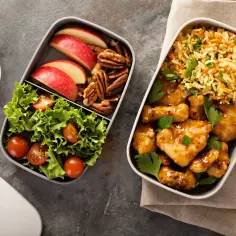 Dwa pojemniki na lunch: jeden z kurczakiem w sosie i ryżem, drugi z sałatką, jabłkiem i orzechami.