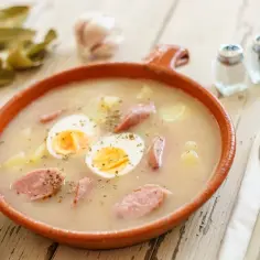 Żurek w glinianej misce z jajkiem, kiełbasą, ziemniakami i pieczywem.