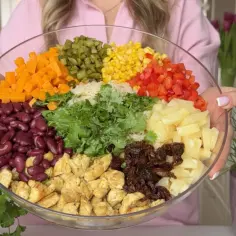 Duża szklana misa z kolorowymi składnikami sałatki: fasola, kurczak, ananas, kukurydza, papryka, marchewka, ogórek, zioła.