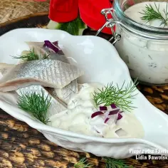 Filety śledziowe w sosie śmietanowym z cebulką i koperkiem, podane w białej miseczce.