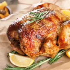 Złocisty kurczak z piekarnika, udekorowany rozmarynem i cytryną, czeka na podanie. Obok miska pieczonych ziemniaków.