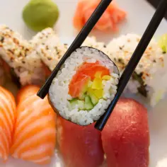 Chopsticks trzymają rolkę sushi z łososiem i warzywami. Na talerzu widać nigiri z łososiem i tuńczykiem, a także inne rolki. Idealna zaprawa do sushi czeka na skosztowanie.