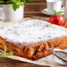 Pyszne, ekspresowe ciasto z jabłkami, posypane cukrem pudrem, obok jabłka i cynamon.