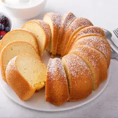 Puszysta babka z cukrem pudrem, pokrojona w plastry, obok miseczka z owocami leśnymi. Idealna na najłatwiejsza babka przepis.
