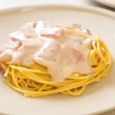 Spaghetti z kremowym sosem śmietanowym i kawałkami szynki na białym talerzu.