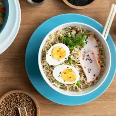 Miseczka tantanmen z makaronem, jajkiem, plastrami mięsa i zielonym groszkiem. Obok sos i przyprawy.