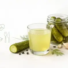 Szklanka wody z ogórków kiszonych, obok słoik z ogórkami, koperek, czosnek i ziarna pieprzu.