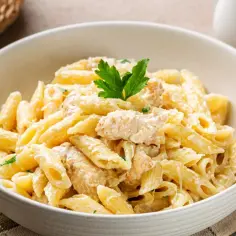 Pyszny makaron penne w kremowym sosie śmietanowym z kawałkami kurczaka, udekorowany natką pietruszki.