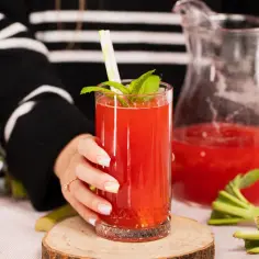 Szklanka domowego kompotu z truskawek i rabarbaru, obok dzbanek, miska truskawek i łodygi rabarbaru.