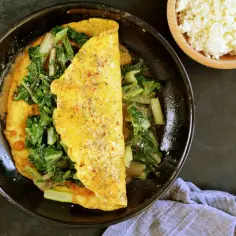 Omlet z serkiem wiejskim i zielonymi warzywami na patelni.
