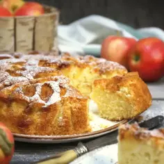 Wilgotne ciasto z jabłkami na oleju, posypane cukrem pudrem, wygląda apetycznie. Obok koszyk z jabłkami i kawałki ciasta.
