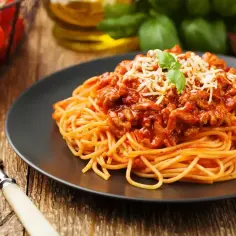 Talerz spaghetti bolognese z serem i bazylią, obok widelec i koszyk pomidorów.