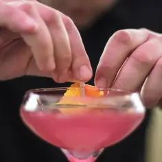 Dłonie dekorują różowy drink skórką pomarańczy.