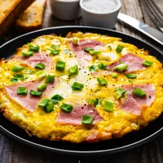 Omlet z szynką i szczypiorkiem na czarnym talerzu, obok grzanki i sztućce.