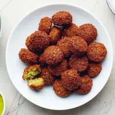 Talerz pełen chrupiących falafeli, jeden przekrojony, obok frytki i sos.