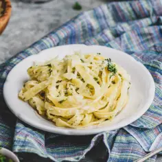 Talerz makaronu tagliatelle z kremowym sosem i startym parmezanem, udekorowany natką pietruszki.
