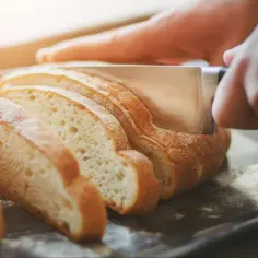 Dłoń kroi nóżem kromki chleba na desce.