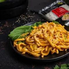 Talerz makaronu udon w sosie seczuańskim, posypany sezamem i z dodatkiem zielonych liści.