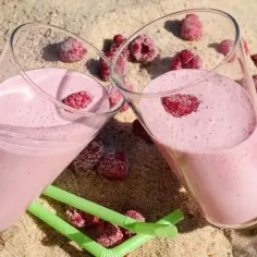 Dwa szklanki różowego smoothie z malinami na piasku, obok zielone słomki i maliny.