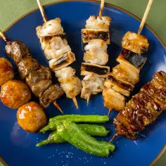 Różnorodne yakitori na talerzu: kurczak, wątróbka, klopsiki i papryczki shishito, polane sosem.