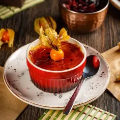 Deser crème brûlée w czerwonej kokilce, ozdobiony physalis, na talerzyku z łyżeczką.