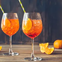 Dwa kieliszki Aperol Spritz z plasterkami pomarańczy i słomkami na drewnianym stole.