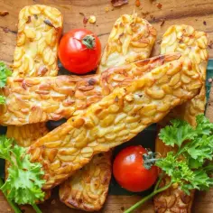 Usmażony tempeh z pomidorkami koktajlowymi i natką pietruszki na drewnianej desce.