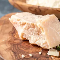 Kawałek twardego sera, przypominającego Grana Padano i parmezan, leży na drewnianej desce. W tle widać misę z tartym serem.