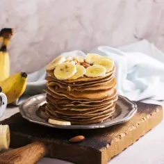Stos naleśników z plasterkami banana i orzechami na talerzu, obok banany i kubek.