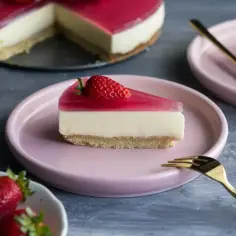 Pyszny deser z mascarpone i galaretką, udekorowany truskawką, na różowym talerzyku. Obok kawałek ciasta i widelczyk.