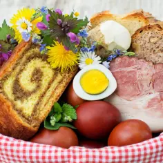 Koszyk wielkanocny z babką, jajkami, chlebem, chrzanem i mięsem, ozdobiony kwiatami.