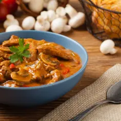 Strogonow wieprzowy z pieczarkami i papryką w niebieskiej misce, udekorowany natką pietruszki.