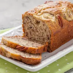 Wilgotny chlebek bananowy z orzechami pekan i plasterkami banana na wierzchu, podany na białej paterze.