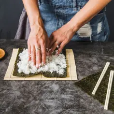 Dłonie rozprowadzają ryż do sushi na nori. Obok leżą pałeczki i marynowany imbir.