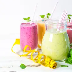 Trzy kolorowe koktajle, dzbanek z zielonym smoothie, miarka krawiecka i listki mięty.