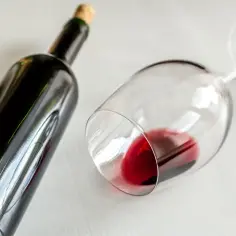 Butelka ciemnego wina i przewrócony kieliszek z czerwonym winem na białym tle.