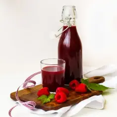 Butelka i szklanka z ciemnoczerwonym sokiem malinowym, maliny i listki mięty na desce.