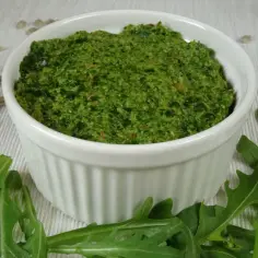 Pesto z rukoli w białej miseczce, otoczone świeżymi liśćmi rukoli i nasionami słonecznika.