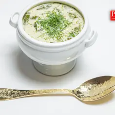 Biała miska z zupą i zielonymi dodatkami, obok złota łyżka z ozdobnym wzorem.