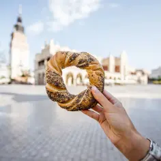 Dłoń trzyma obwarzanka krakowskiego na tle Sukiennic.