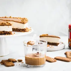 Sernik z kremem Biscoff i pokruszonymi ciastkami Lotus. Słoik kremu Biscoff i kilka ciastek na blacie.