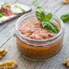 Słoiczek z domowym, czerwonym pesto, ozdobiony listkiem bazylii. Obok orzechy włoskie i kromka chleba z serkiem.