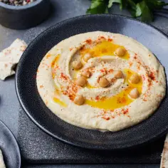 Kremowy hummus z ciecierzycą, oliwą i papryką na czarnym talerzu, obok pity i czosnku.