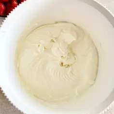 Kremowy serek mascarpone w białej misie, gotowy do deseru. Obok miski widać miseczkę z truskawkami.
