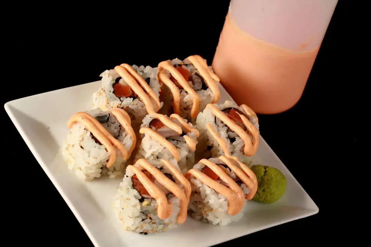 Roladki sushi polane kremowym, pomarańczowym sosem. Obok porcja wasabi i butelka z resztą sosu do sushi.