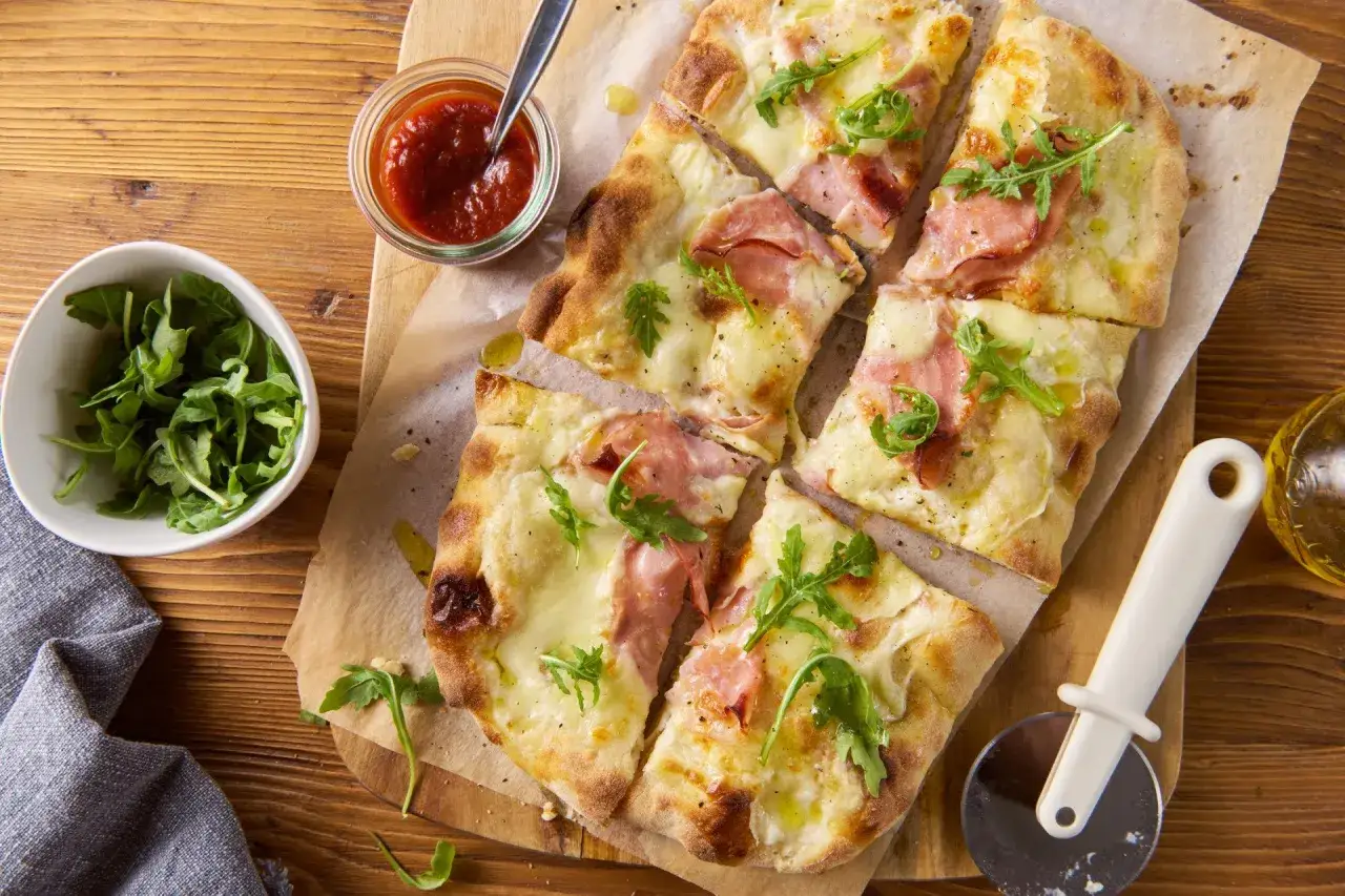 Pyszna pizza bianca z szynką, mozzarellą i rukolą, podana z sosem pomidorowym i oliwą. Idealna na szybki lunch.