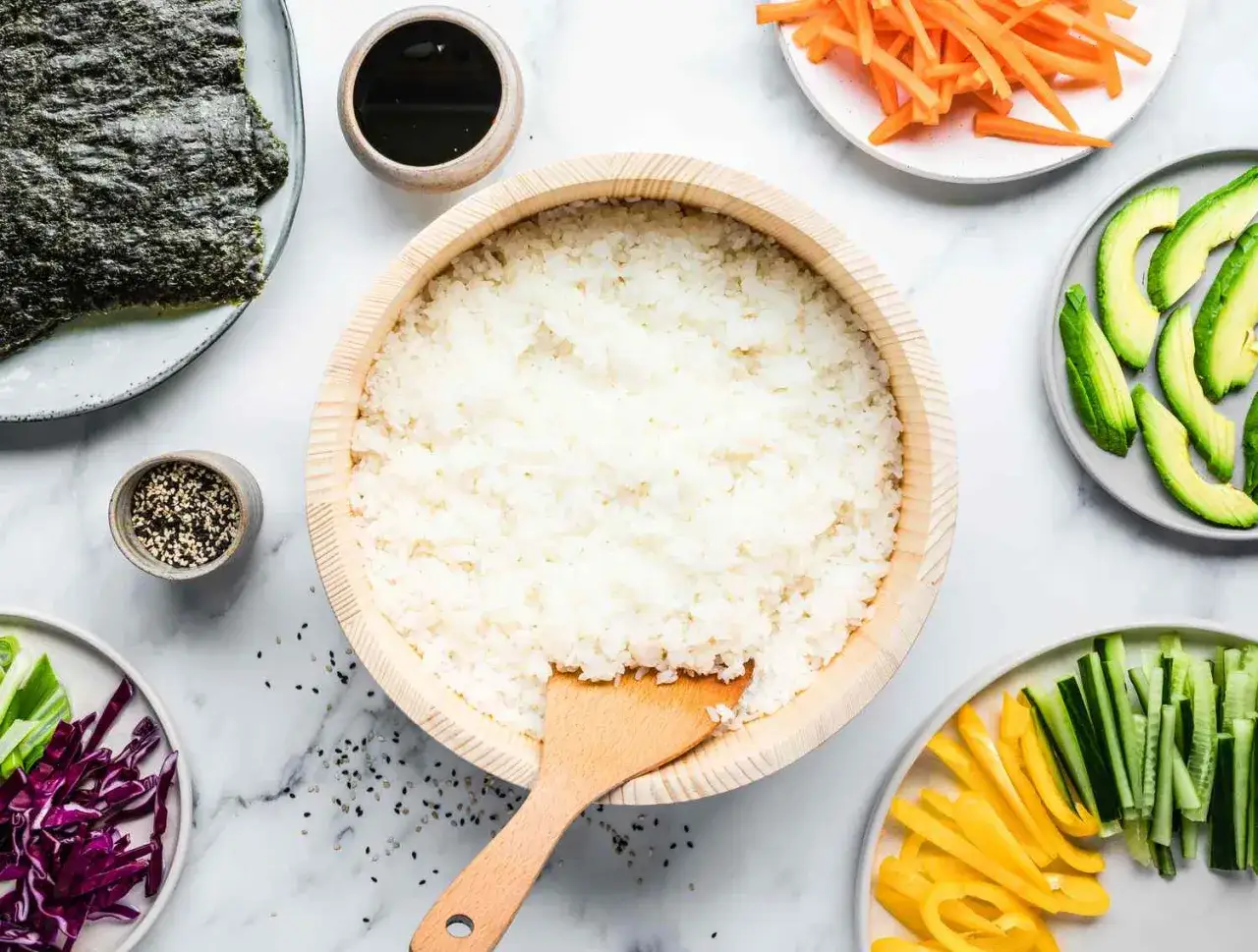 Gotowy ryż do sushi w drewnianej misie, obok nori, awokado, marchewka, ogórek, papryka, cebulka i sezam.