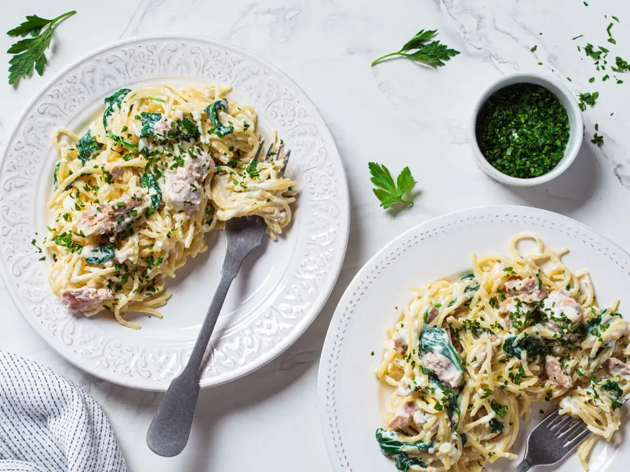 Dwa talerze spaghetti z kremowym sosem, szpinakiem i kawałkami tuńczyka z puszki, posypane natką pietruszki.